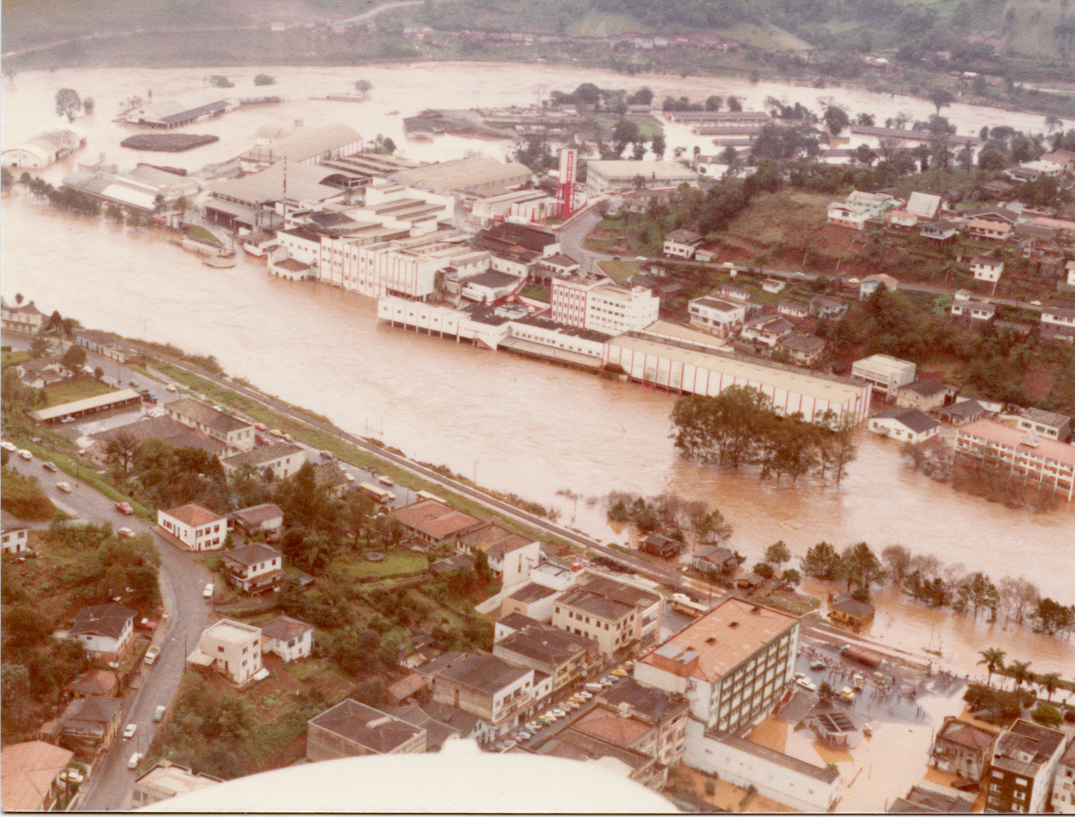 Uma fotografia aérea que registra uma enchente severa em uma área urbana, possivelmente em uma cidade do interior catarinense como Videira. O rio transbordou, inundando ruas e atingindo a base de grandes complexos industriais e residências, com a água barrenta cobrindo grande parte da paisagem.