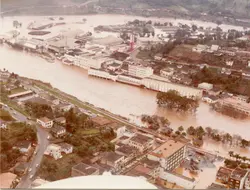 Uma fotografia aérea que registra uma enchente severa em uma área urbana, possivelmente em uma cidade do interior catarinense como Videira. O rio transbordou, inundando ruas e atingindo a base de grandes complexos industriais e residências, com a água barrenta cobrindo grande parte da paisagem.