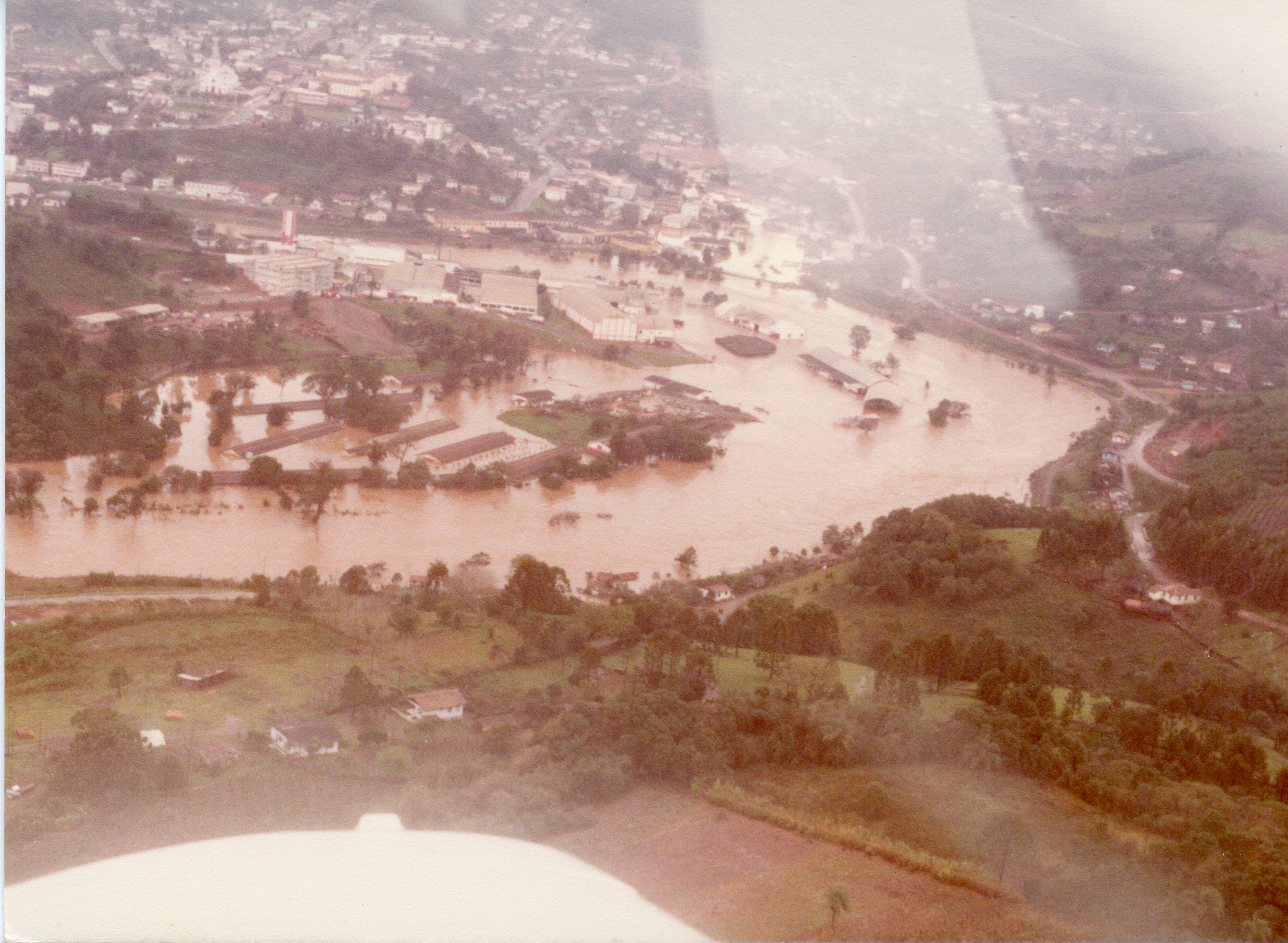 Fotografia aérea mostrando uma enchente severa em uma área urbana e industrial, possivelmente em Videira, Santa Catarina. A imagem captura o transbordamento de um rio que inunda galpões e estruturas próximas, com o restante da cidade visível ao fundo, sugerindo um evento climático significativo ocorrido provavelmente entre as décadas de 1970 e 1980.