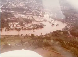 Fotografia aérea mostrando uma enchente severa em uma área urbana e industrial, possivelmente em Videira, Santa Catarina. A imagem captura o transbordamento de um rio que inunda galpões e estruturas próximas, com o restante da cidade visível ao fundo, sugerindo um evento climático significativo ocorrido provavelmente entre as décadas de 1970 e 1980.