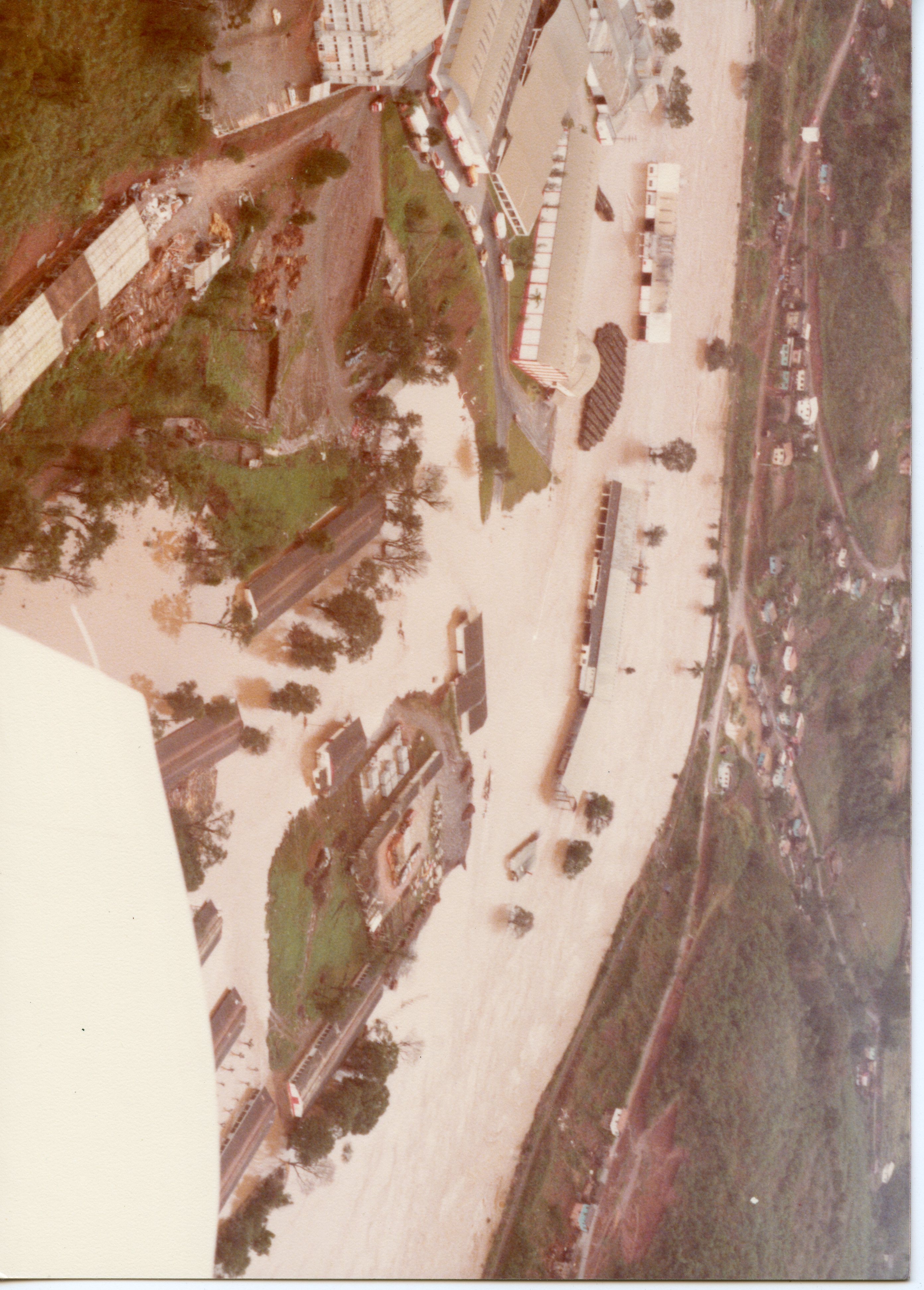 Vista aérea de uma área industrial ou urbana possivelmente afetada por uma enchente, com grandes extensões de terreno cobertas por sedimentos ou água barrenta. A imagem mostra galpões, estruturas fabris e uma topografia típica de regiões do interior do Brasil, sugerindo um registro documental de um evento climático ou de expansão industrial.