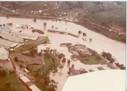 Vista aérea de uma área industrial ou urbana possivelmente afetada por uma enchente, com grandes extensões de terreno cobertas por sedimentos ou água barrenta. A imagem mostra galpões, estruturas fabris e uma topografia típica de regiões do interior do Brasil, sugerindo um registro documental de um evento climático ou de expansão industrial.