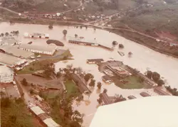 Vista aérea de uma área industrial ou urbana afetada por uma enchente severa, com águas barrentas cobrindo grande parte do terreno e cercando construções. A imagem, provavelmente da segunda metade do século XX, mostra galpões e infraestrutura logística submersos, sugerindo um evento climático de grande impacto em uma região brasileira.