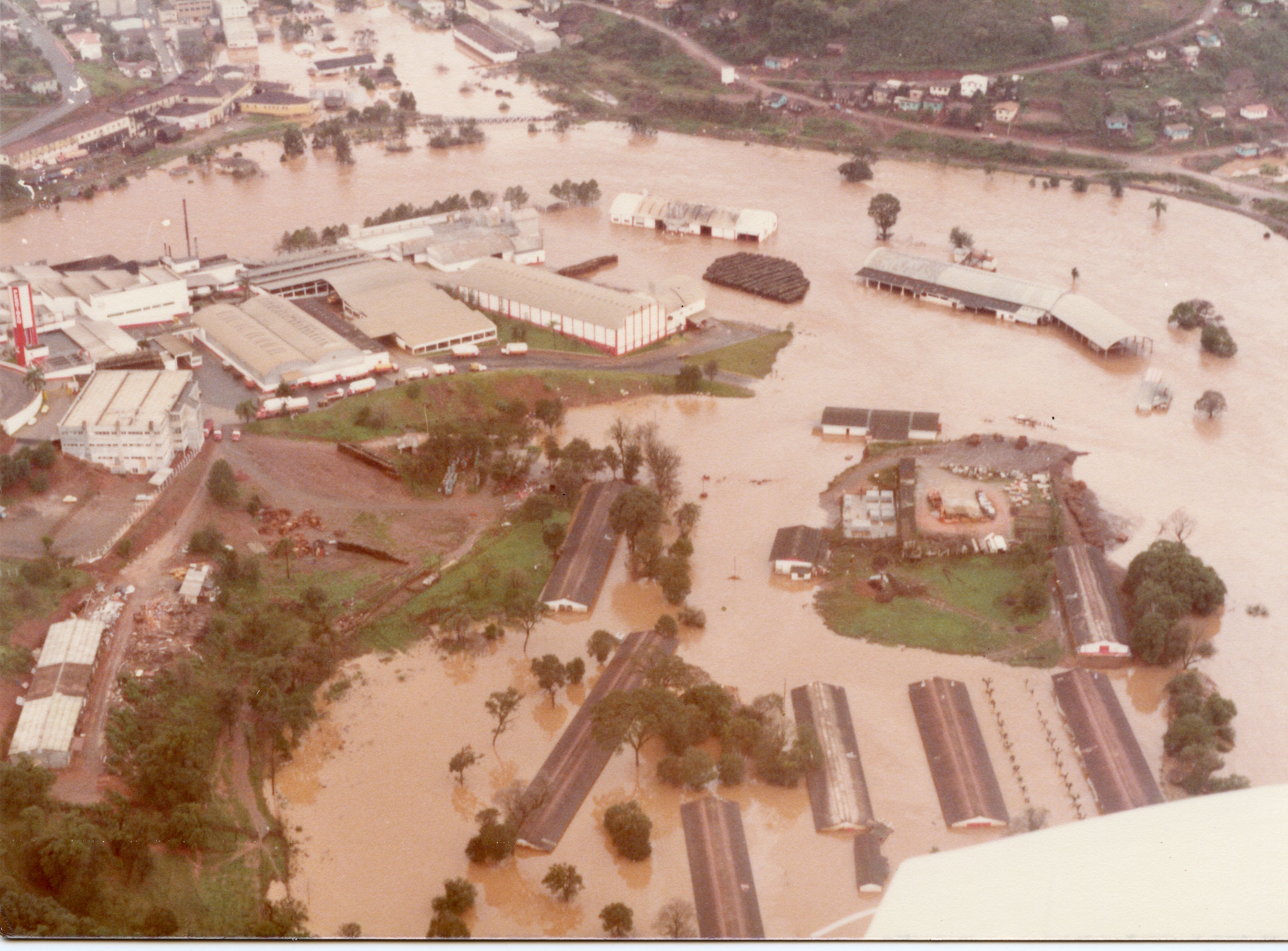 Uma vista aérea de uma enchente severa em uma área industrial e urbana, possivelmente em Videira, Santa Catarina. A imagem mostra galpões e estruturas parcialmente submersos por águas barrentas, sugerindo um evento climático significativo ocorrido provavelmente nas décadas de 1980 ou 1990.