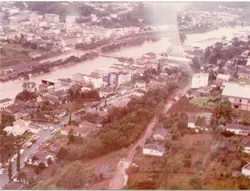 Uma fotografia aérea de uma cidade brasileira, possivelmente Videira, mostrando uma enchente que cobre parte da área urbana e margens do rio. A imagem apresenta tons desbotados típicos de fotografias coloridas das décadas de 1970 ou 1980, destacando a infraestrutura urbana e a vegetação ao redor.