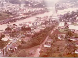 Uma vista aérea de uma cidade brasileira, possivelmente Videira, durante um evento de enchente que inundou áreas próximas ao rio. A imagem mostra o tecido urbano com ruas, casas e edifícios comerciais, com a água barrenta ocupando parte significativa da paisagem, sugerindo um desastre natural ocorrido provavelmente entre as décadas de 1970 e 1980.