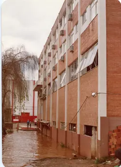 Uma fotografia que registra uma enchente urbana, mostrando o nível da água atingindo a base de um edifício de tijolos aparentes com várias janelas. A cena sugere um evento climático em uma cidade brasileira, possivelmente nas décadas de 1980 ou 1990, com pessoas observando a situação ao fundo.