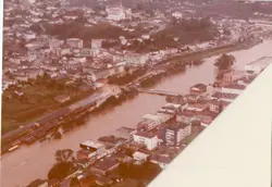 Uma fotografia aérea de uma cidade, possivelmente Videira, mostrando um rio com águas barrentas que parece ter transbordado ou atingido um nível elevado. A imagem apresenta uma perspectiva urbana com edifícios e ruas, sugerindo um evento de enchente ocorrido provavelmente entre as décadas de 1970 e 1980.