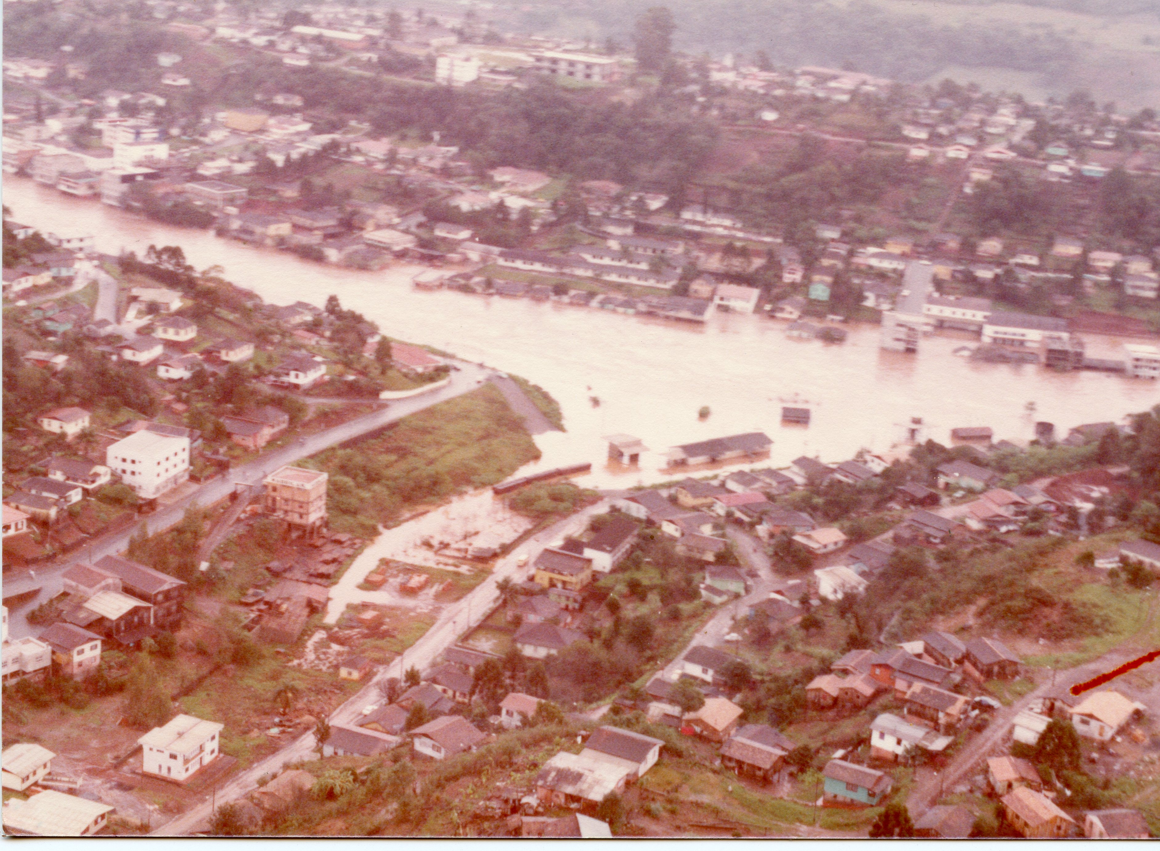 Uma vista aérea de uma enchente em uma pequena cidade brasileira, mostrando o rio transbordado atingindo casas e ruas. A imagem apresenta tons desbotados típicos de fotografias coloridas das décadas de 1970 ou 1980, capturando um evento climático significativo que impactou a infraestrutura urbana local.