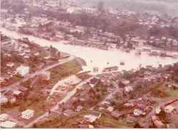 Uma vista aérea de uma enchente em uma pequena cidade brasileira, mostrando o rio transbordado atingindo casas e ruas. A imagem apresenta tons desbotados típicos de fotografias coloridas das décadas de 1970 ou 1980, capturando um evento climático significativo que impactou a infraestrutura urbana local.