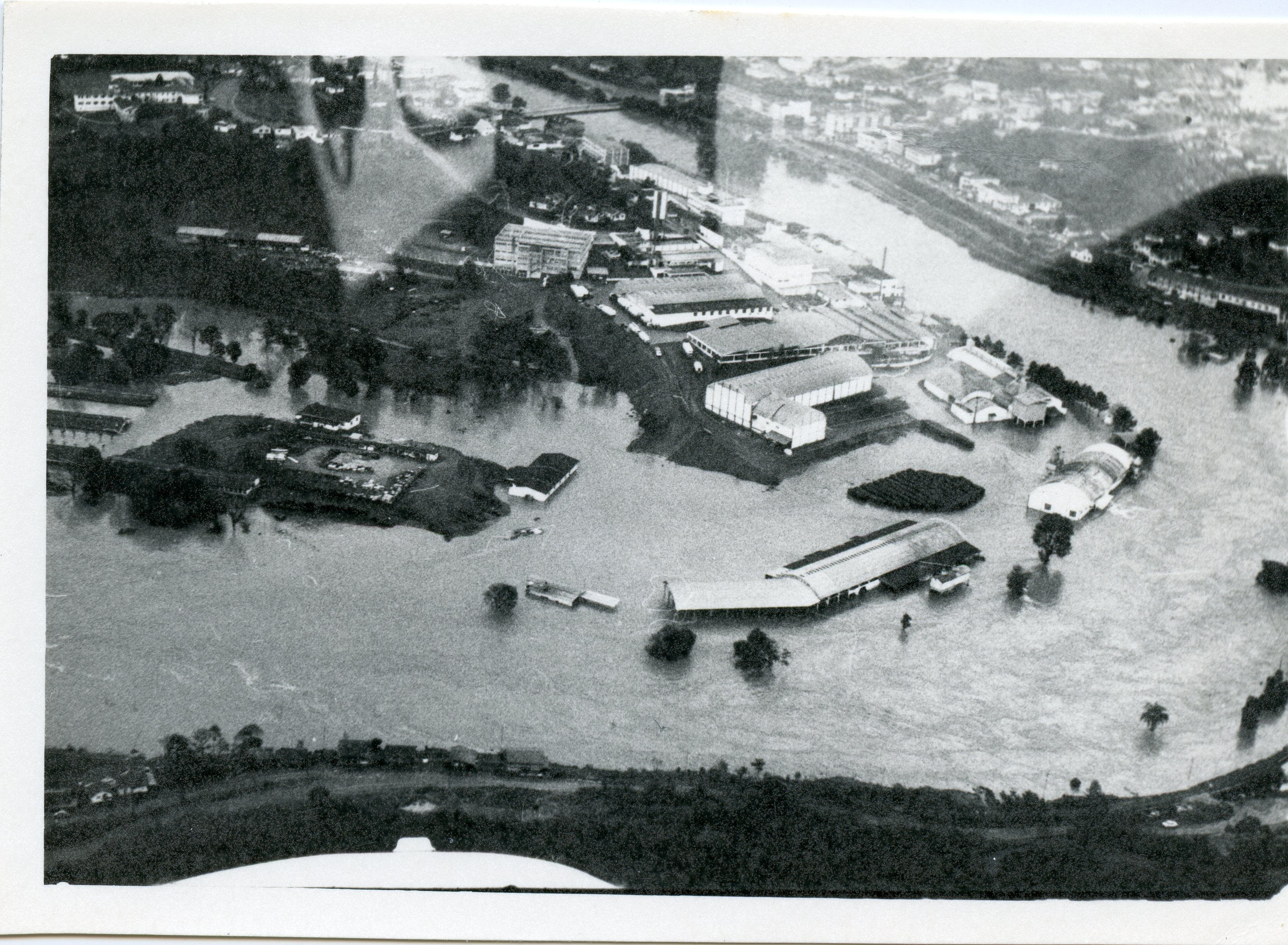 Fotografia aérea em preto e branco registrando uma enchente severa em uma área urbana industrial, possivelmente em uma cidade do interior catarinense como Videira. A imagem mostra galpões e construções parcialmente submersos pelas águas de um rio que transbordou, evidenciando um evento climático de grande impacto na infraestrutura local.
