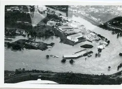 Fotografia aérea em preto e branco registrando uma enchente severa em uma área urbana industrial, possivelmente em uma cidade do interior catarinense como Videira. A imagem mostra galpões e construções parcialmente submersos pelas águas de um rio que transbordou, evidenciando um evento climático de grande impacto na infraestrutura local.
