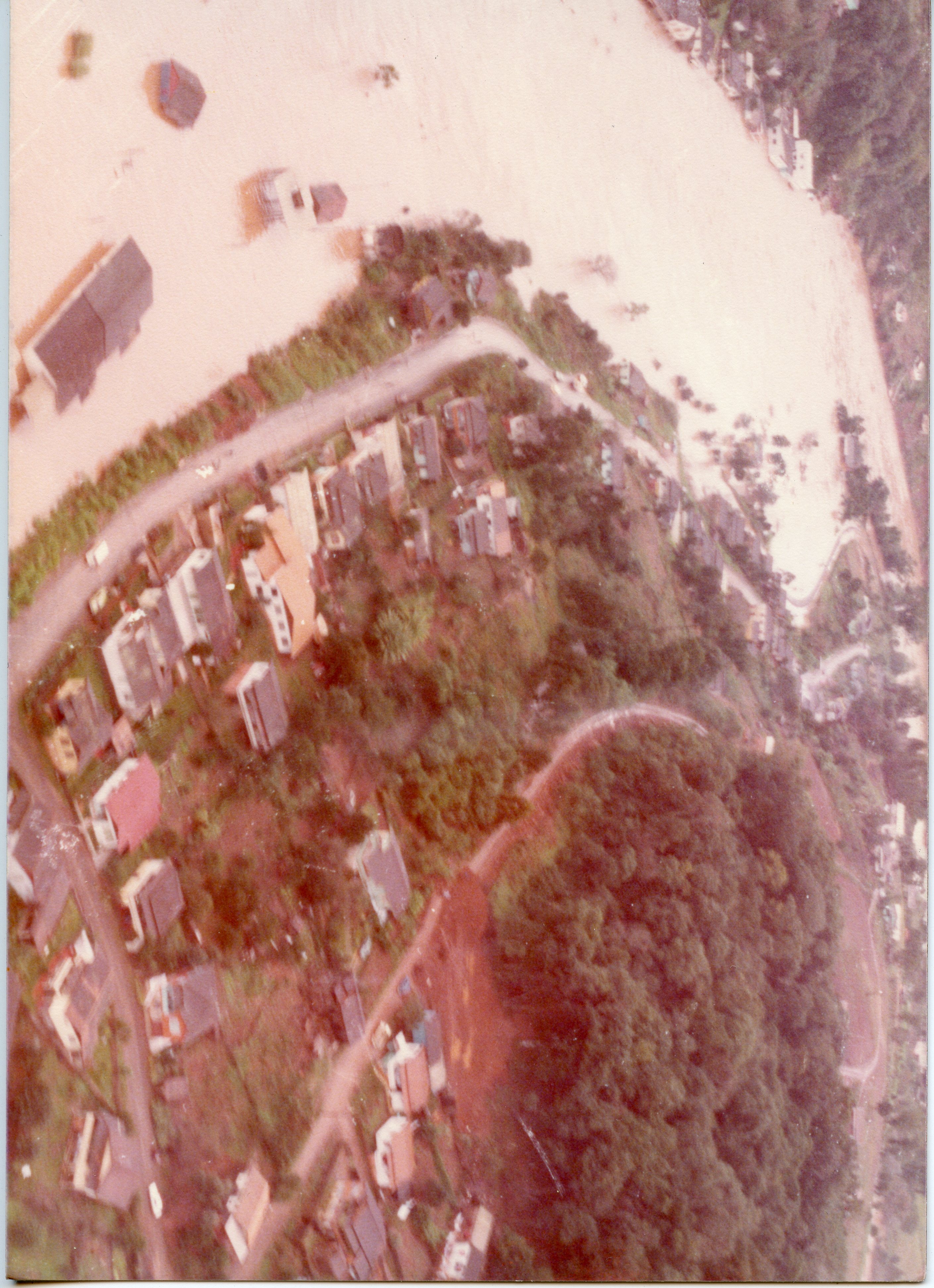 Fotografia aérea de uma enchente em uma área urbana, possivelmente em uma cidade do interior brasileiro. A imagem mostra casas parcialmente submersas pelas águas barrentas de um rio, com vegetação e estradas visíveis, sugerindo um evento climático severo ocorrido provavelmente entre as décadas de 1970 e 1980.
