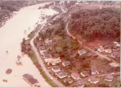 Fotografia aérea de uma enchente em uma área urbana, possivelmente em uma cidade do interior brasileiro. A imagem mostra casas parcialmente submersas pelas águas barrentas de um rio, com vegetação e estradas visíveis, sugerindo um evento climático severo ocorrido provavelmente entre as décadas de 1970 e 1980.