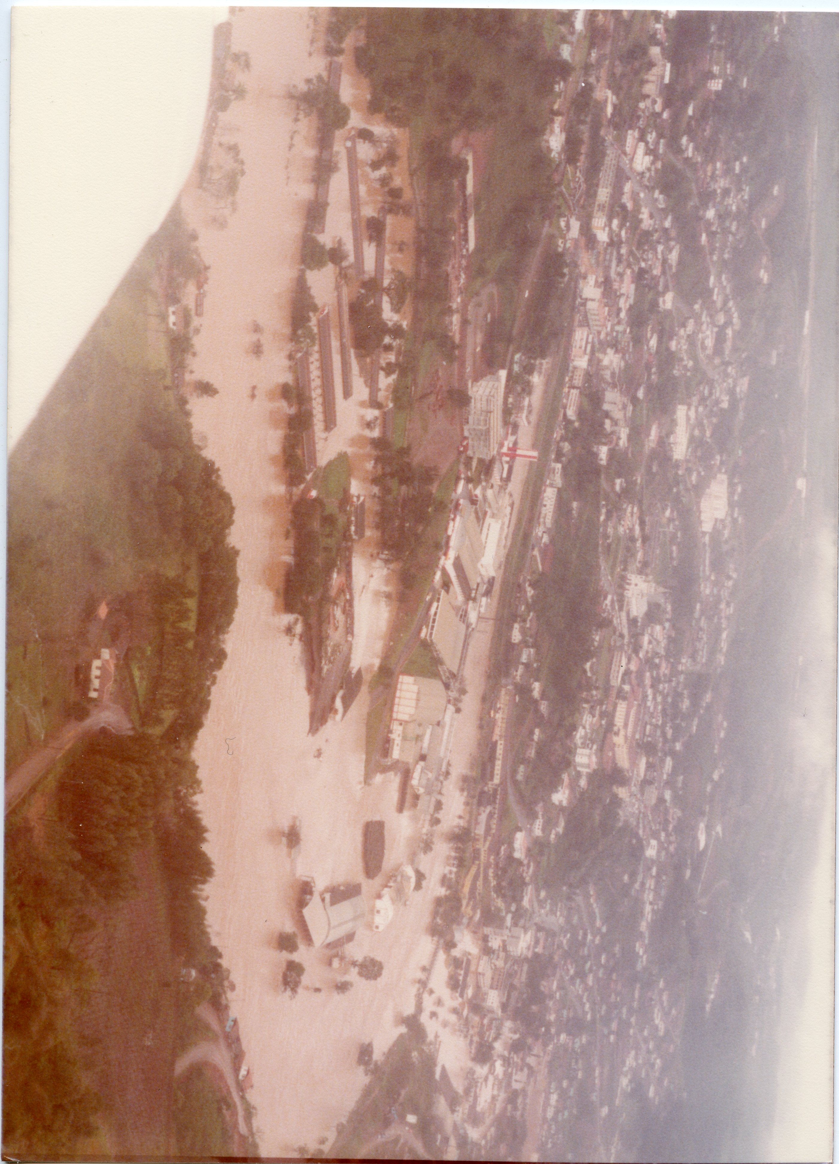 Fotografia aérea mostrando uma enchente severa que inunda áreas urbanas e industriais próximas a um rio. A imagem registra um evento climático significativo, possivelmente em uma cidade do interior catarinense, com construções parcialmente submersas e sedimentos visíveis na água.
