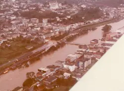 Vista aérea de uma enchente em uma cidade brasileira, possivelmente Videira, com o rio transbordado atingindo áreas urbanas e ferrovias. A fotografia apresenta tons sépia característicos de registros fotográficos das décadas de 1970 ou 1980, mostrando a infraestrutura urbana parcialmente submersa.