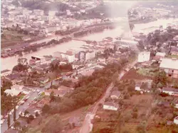 Vista aérea de uma cidade brasileira, possivelmente Videira, durante um evento de enchente que cobre parte das margens do rio. A imagem apresenta arquitetura urbana típica de meados do século XX, com ruas pavimentadas e vegetação densa nas encostas.