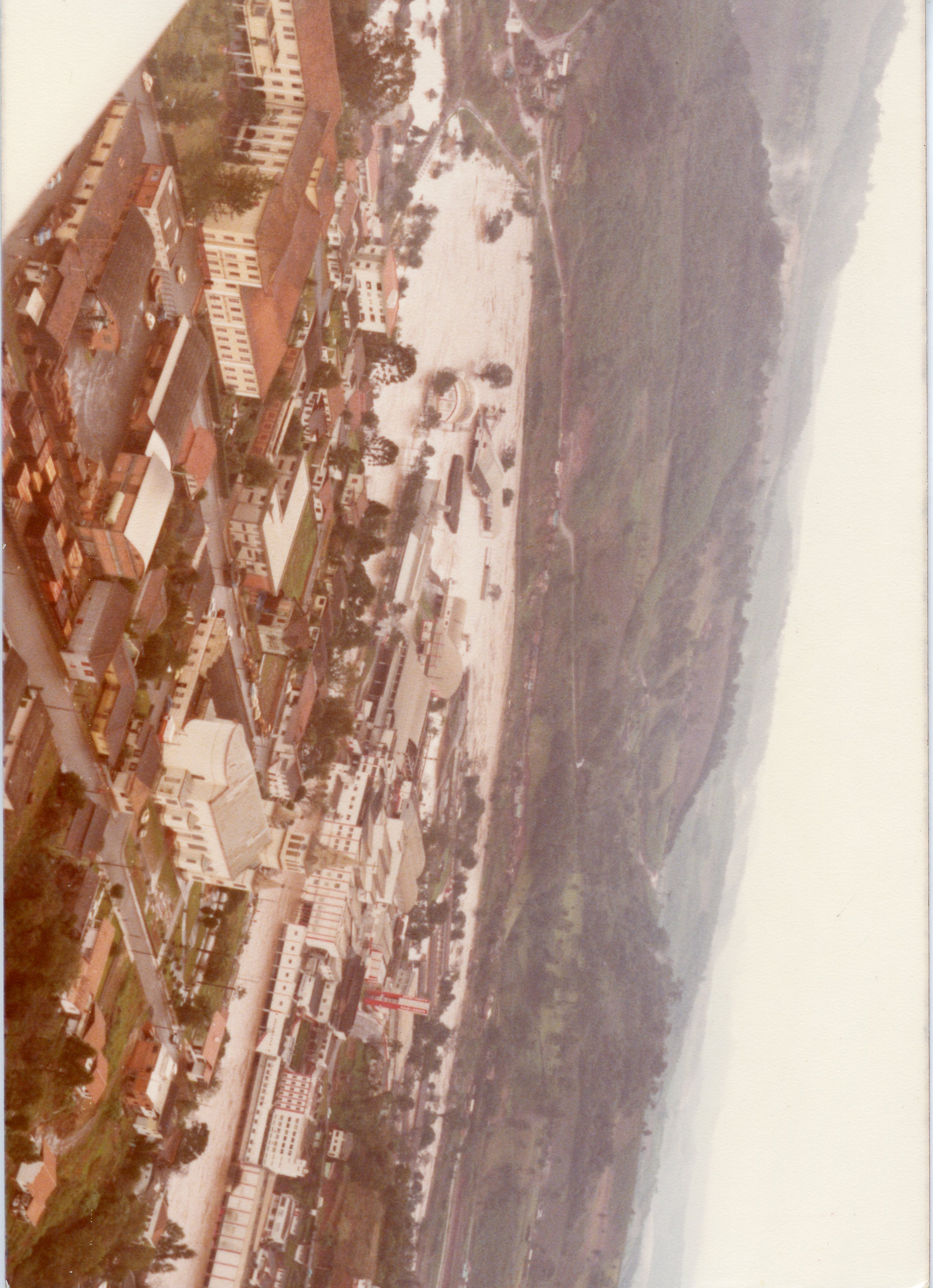 Uma fotografia aérea de uma pequena cidade brasileira, possivelmente Videira, mostrando uma área urbana densa próxima a uma encosta e uma grande área alagada ou inundada. A imagem apresenta tons desbotados típicos de fotografias coloridas das décadas de 1970 ou 1980, destacando a topografia acidentada e o planejamento urbano inicial.