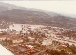 Uma fotografia aérea de uma pequena cidade brasileira, possivelmente Videira, mostrando uma área urbana densa próxima a uma encosta e uma grande área alagada ou inundada. A imagem apresenta tons desbotados típicos de fotografias coloridas das décadas de 1970 ou 1980, destacando a topografia acidentada e o planejamento urbano inicial.