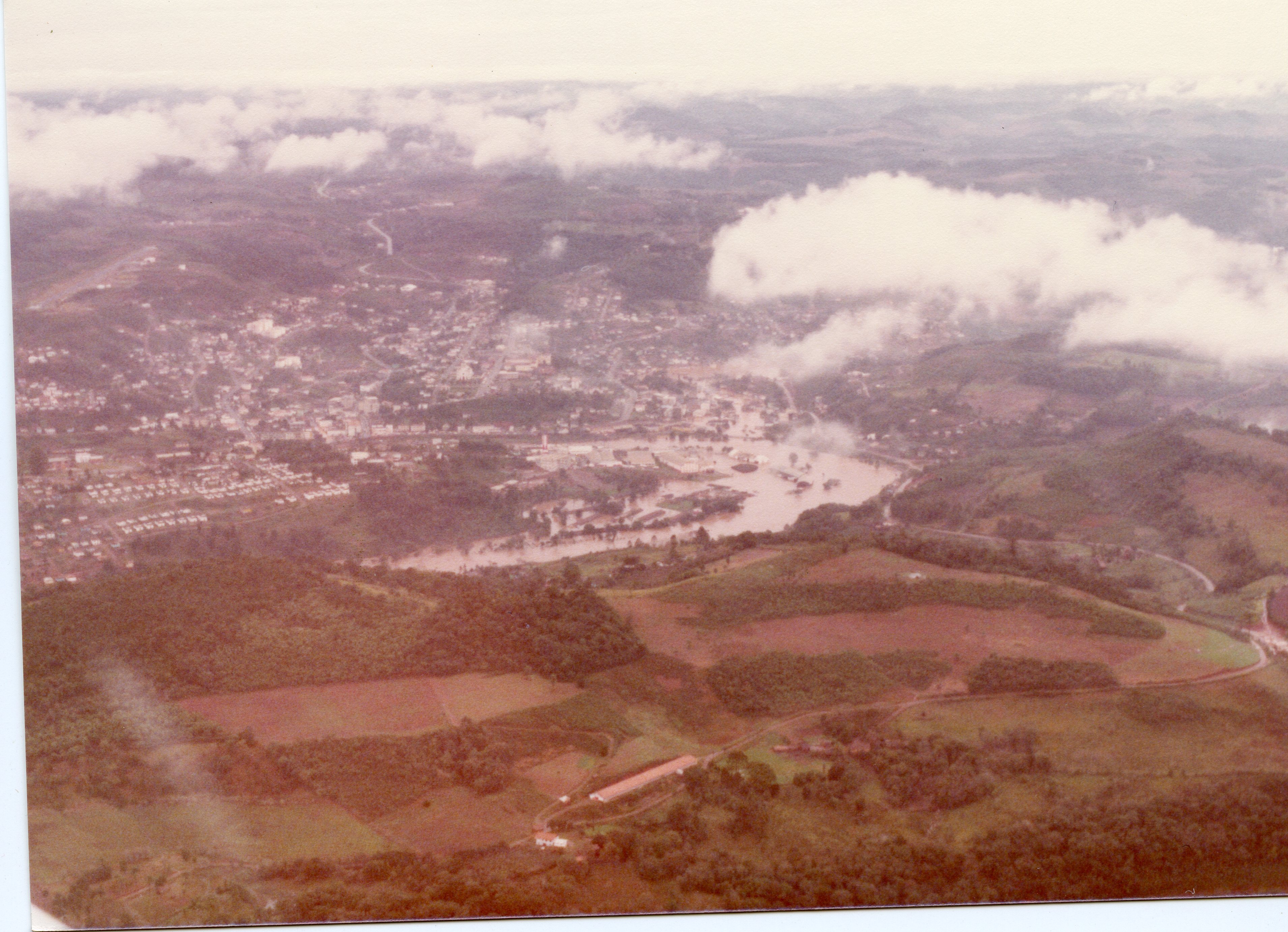 Uma vista aérea panorâmica de uma cidade brasileira, possivelmente Videira, mostrando uma área urbana densa cortada por um rio e cercada por colinas e vegetação. A imagem apresenta tons desbotados típicos de fotografias coloridas das décadas de 1970 ou 1980, capturando o desenvolvimento urbano e a topografia local.