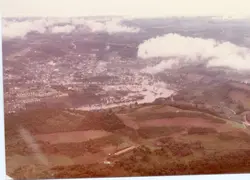 Uma vista aérea panorâmica de uma cidade brasileira, possivelmente Videira, mostrando uma área urbana densa cortada por um rio e cercada por colinas e vegetação. A imagem apresenta tons desbotados típicos de fotografias coloridas das décadas de 1970 ou 1980, capturando o desenvolvimento urbano e a topografia local.