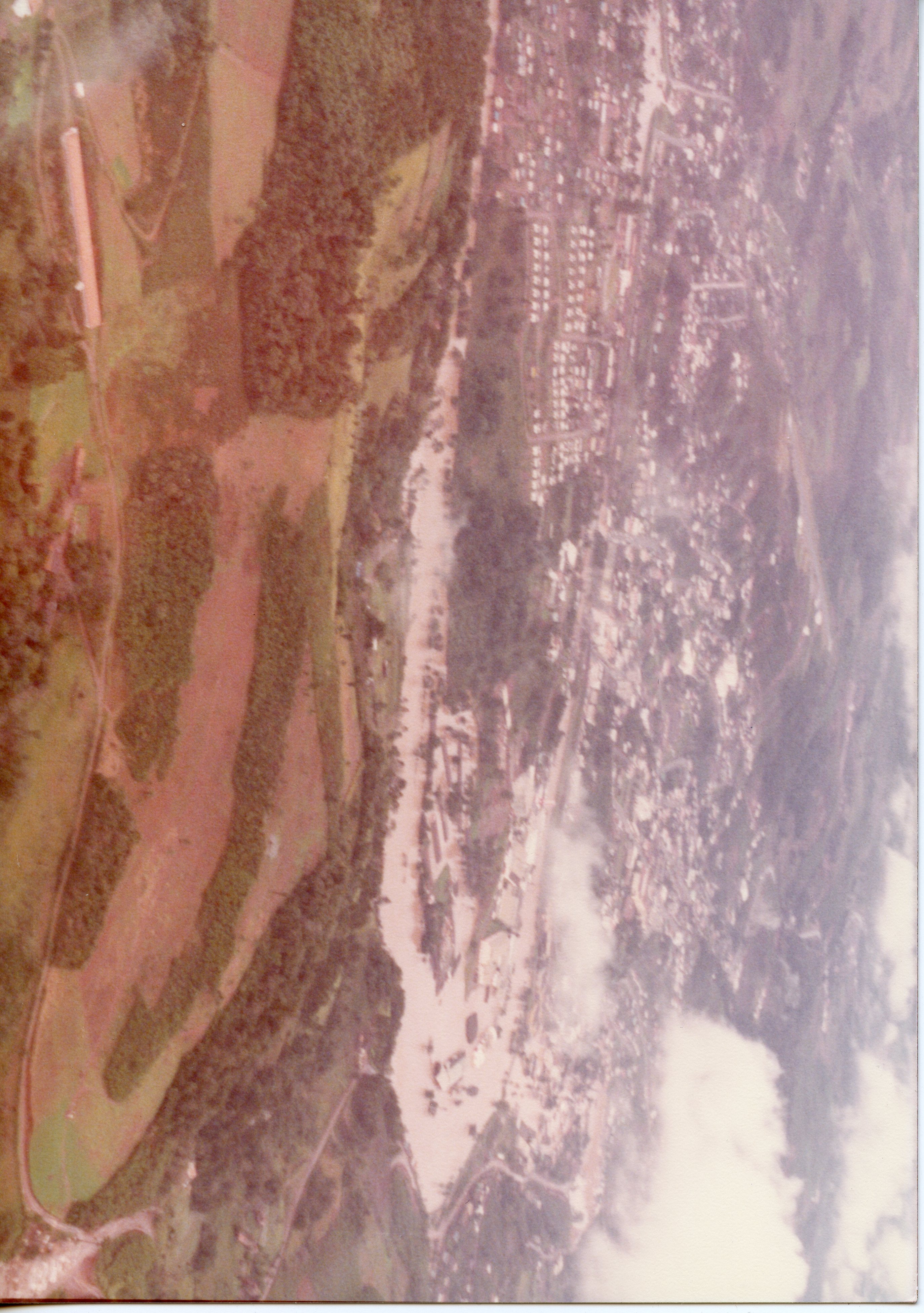 Uma vista aérea de uma pequena cidade brasileira, possivelmente Videira, mostrando a ocupação urbana ao lado de áreas rurais e vegetação. A imagem apresenta características de fotografia colorida antiga, com tons desbotados, sugerindo um registro das décadas de 1970 ou 1980.