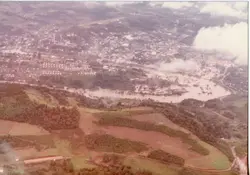 Uma vista aérea de uma pequena cidade brasileira, possivelmente Videira, mostrando a ocupação urbana ao lado de áreas rurais e vegetação. A imagem apresenta características de fotografia colorida antiga, com tons desbotados, sugerindo um registro das décadas de 1970 ou 1980.