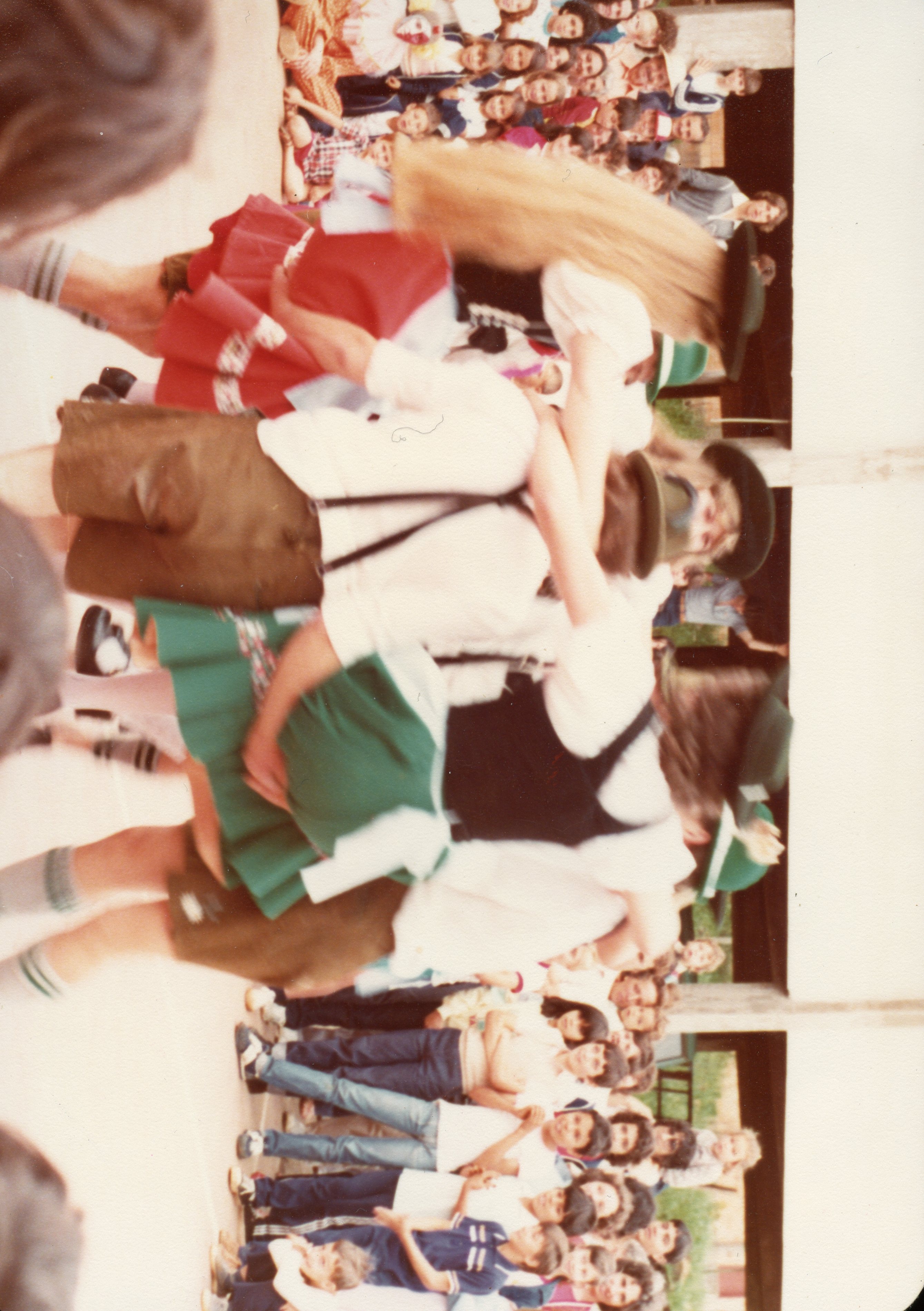Uma fotografia colorida que captura um grupo de pessoas, possivelmente em um evento cultural ou festividade folclórica, com dançarinos em primeiro plano vestindo trajes típicos germânicos. O contexto sugere uma celebração comunitária, comum em regiões de colonização europeia no sul do Brasil, datada provavelmente da década de 1980 devido à qualidade da película e vestimentas.