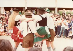 Uma fotografia colorida que captura um grupo de pessoas, possivelmente em um evento cultural ou festividade folclórica, com dançarinos em primeiro plano vestindo trajes típicos germânicos. O contexto sugere uma celebração comunitária, comum em regiões de colonização europeia no sul do Brasil, datada provavelmente da década de 1980 devido à qualidade da película e vestimentas.