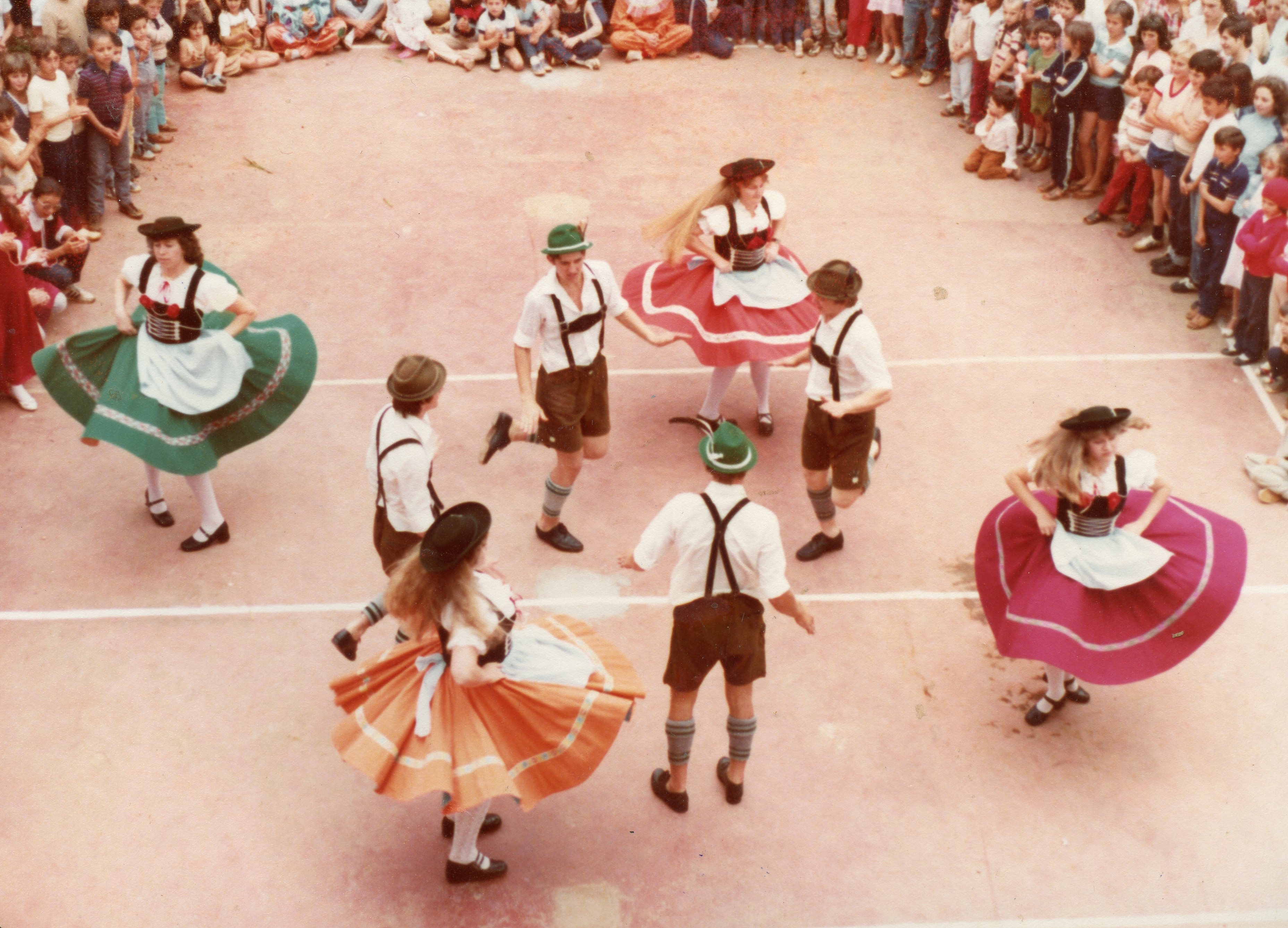 Uma fotografia capturando um grupo de jovens dançando uma dança folclórica de influência alemã, possivelmente em um evento escolar ou cultural no Brasil. Os dançarinos vestem trajes típicos, como lederhosen e saias rodadas, enquanto uma plateia de crianças e adolescentes observa ao redor em uma quadra esportiva.