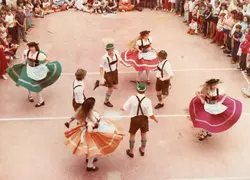 Uma fotografia capturando um grupo de jovens dançando uma dança folclórica de influência alemã, possivelmente em um evento escolar ou cultural no Brasil. Os dançarinos vestem trajes típicos, como lederhosen e saias rodadas, enquanto uma plateia de crianças e adolescentes observa ao redor em uma quadra esportiva.