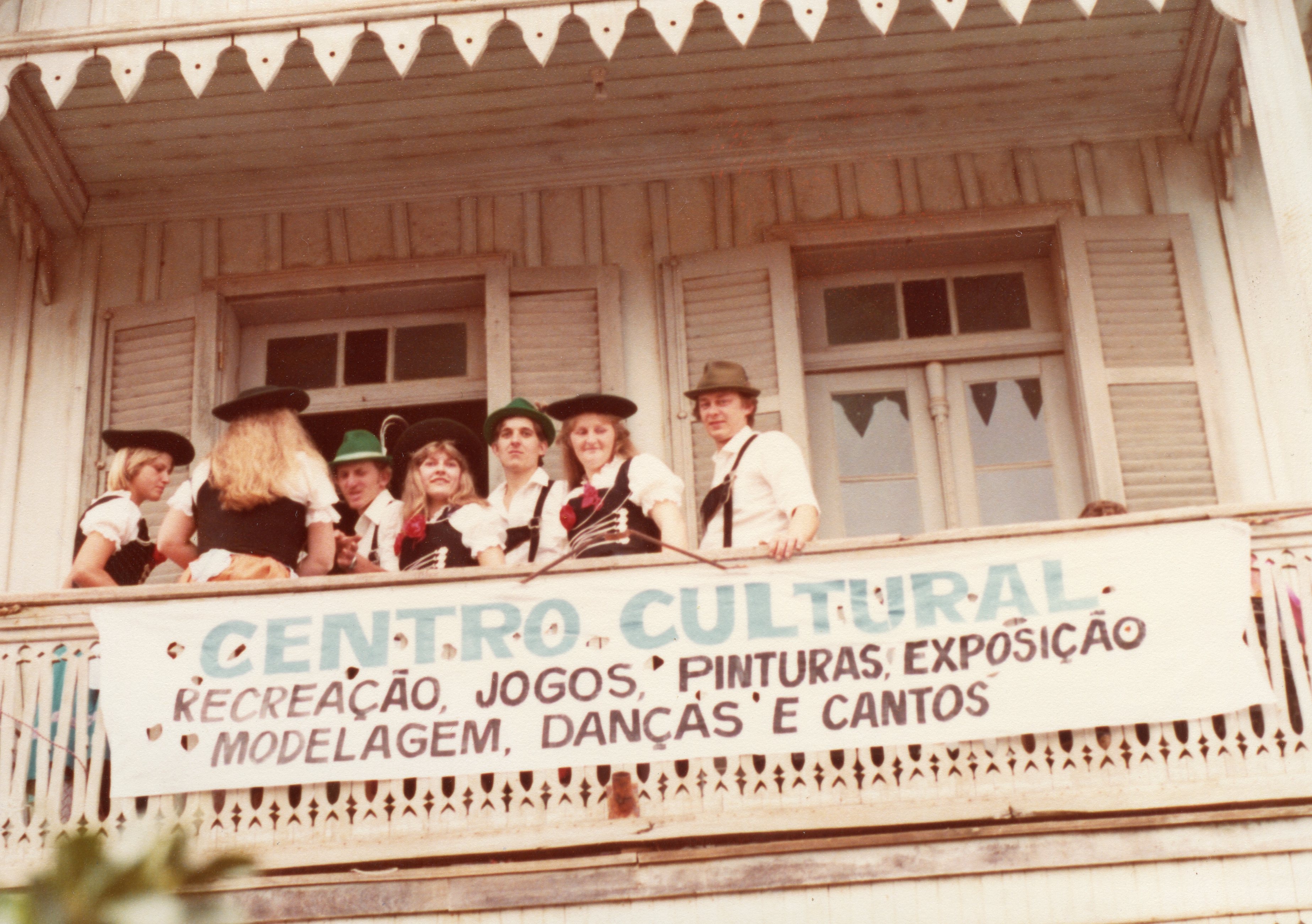 Um grupo de jovens vestidos com trajes típicos alemães posa em uma sacada de madeira de uma construção histórica. A cena sugere um evento de celebração cultural, possivelmente em uma comunidade de colonização europeia no sul do Brasil, com uma faixa informativa sobre atividades culturais.