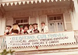 Um grupo de jovens vestidos com trajes típicos alemães posa em uma sacada de madeira de uma construção histórica. A cena sugere um evento de celebração cultural, possivelmente em uma comunidade de colonização europeia no sul do Brasil, com uma faixa informativa sobre atividades culturais.