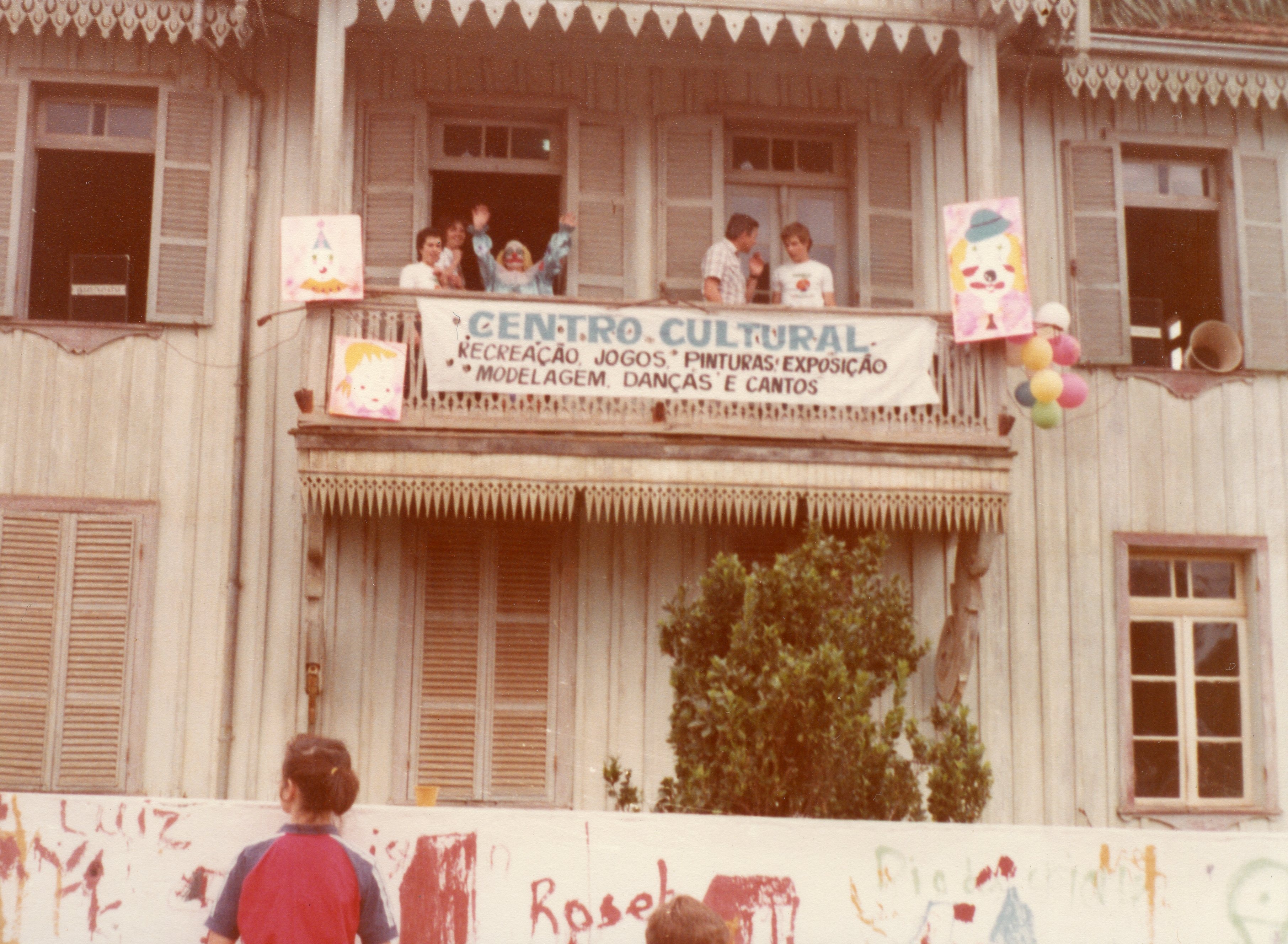 Uma fotografia colorida de época mostrando um casarão de madeira com arquitetura típica de colonização, onde ocorre um evento do Centro Cultural. Pessoas estão na sacada, incluindo um palhaço, com faixas e decorações infantis, sugerindo uma atividade recreativa ou cultural em uma pequena cidade brasileira, provavelmente nas décadas de 1970 ou 1980.