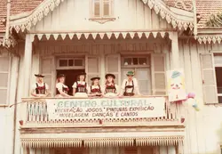 Uma fotografia colorida mostrando um grupo de jovens vestidas com trajes típicos germânicos em uma sacada de madeira de uma casa com arquitetura colonial. O grupo está posicionado atrás de uma faixa que identifica um 'Centro Cultural', sugerindo um evento de preservação da cultura imigrante, possivelmente na região sul do Brasil.