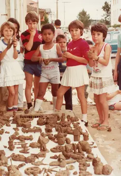Uma fotografia colorida que retrata um grupo de crianças brasileiras brincando com esculturas de barro em uma rua, provavelmente durante uma atividade escolar ou recreativa. As vestimentas, incluindo a camisa do Flamengo e o estilo das saias, sugerem um contexto social e cultural típico do Brasil na década de 1980.