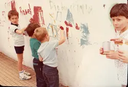 Uma fotografia colorida que captura um momento de lazer infantil, onde crianças pintam uma parede branca com desenhos e escritas. A imagem reflete uma cena cotidiana, possivelmente em um ambiente escolar ou recreativo, com vestimentas típicas das décadas de 1980 ou 1990.