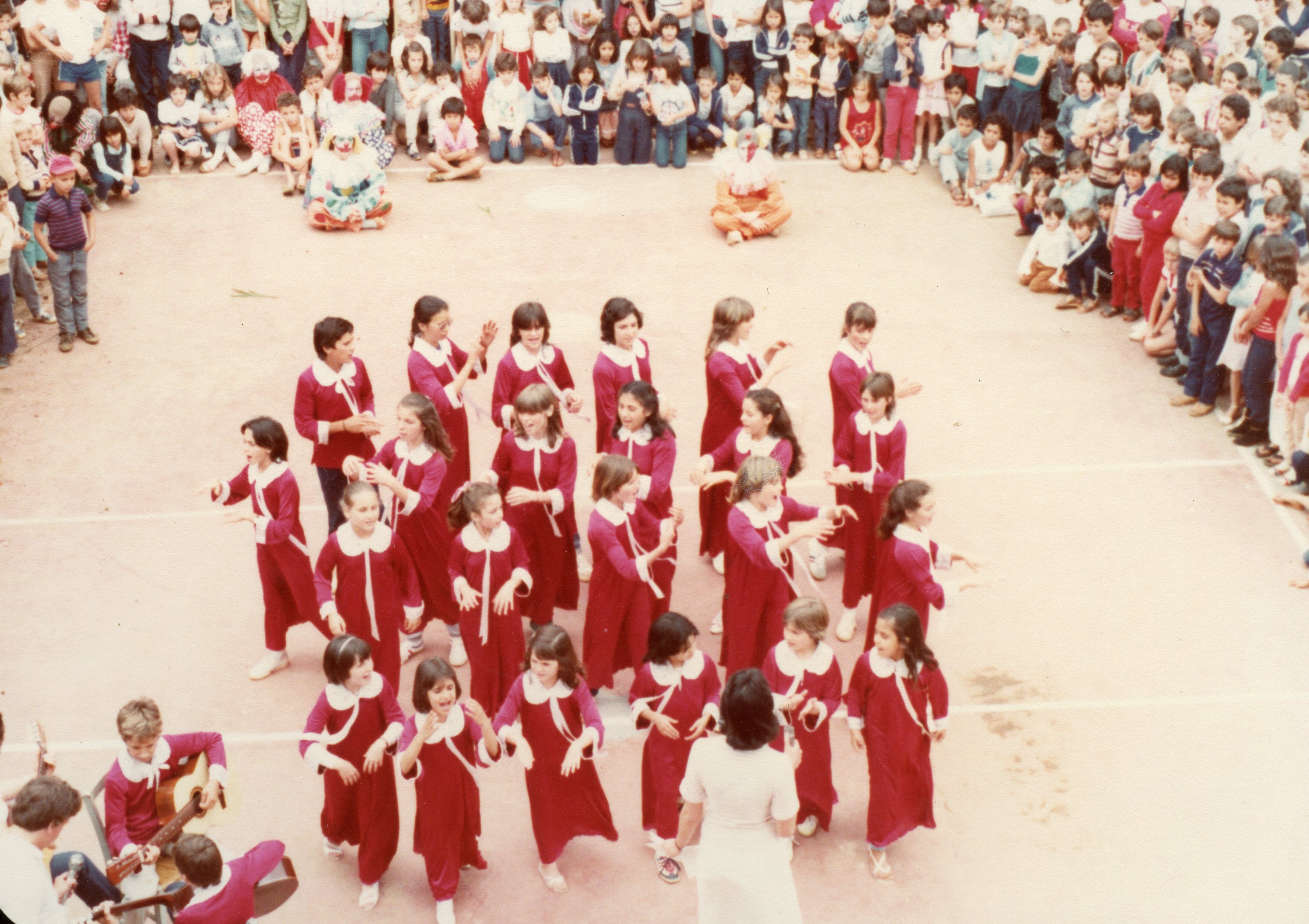 Uma fotografia de um evento escolar ou comunitário ao ar livre, possivelmente na década de 1980, mostrando um grupo de crianças vestidas com túnicas vermelhas e golas brancas realizando uma apresentação. Ao fundo, uma grande plateia de crianças e adultos observa a performance, que conta com a presença de palhaços sentados no chão e um músico tocando violão.