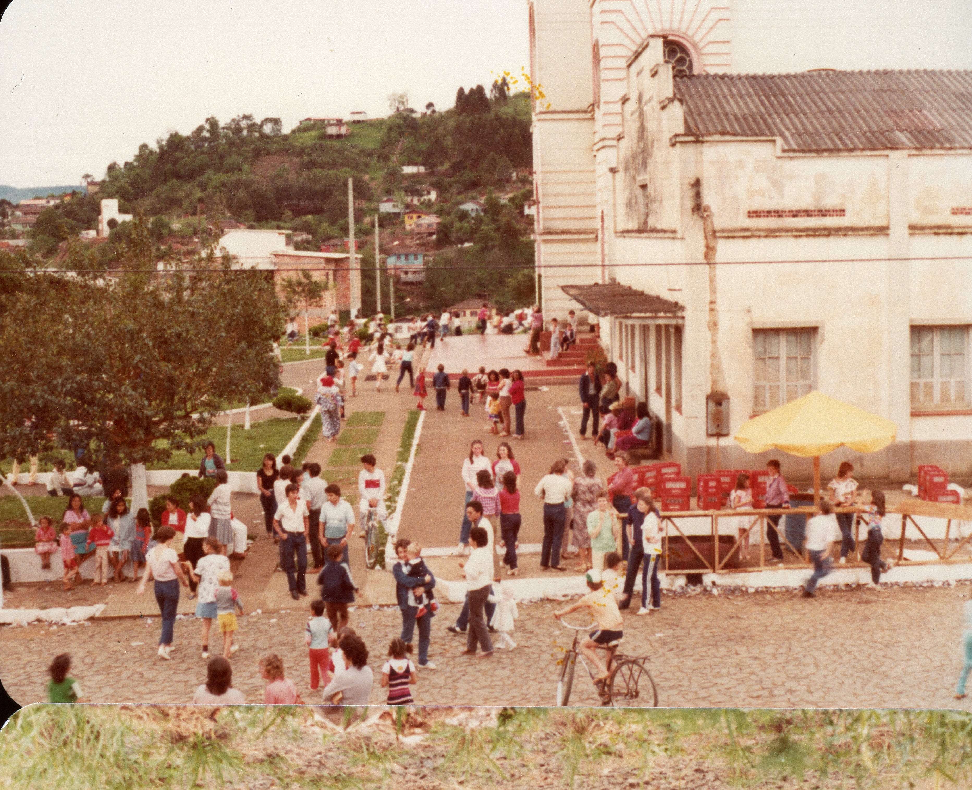 Uma cena ao ar livre em uma pequena cidade brasileira, mostrando uma praça ou pátio adjacente a uma igreja de arquitetura clássica. O ambiente sugere um evento comunitário ou festividade religiosa, com muitas pessoas circulando, crianças brincando e uma estrutura improvisada com guarda-sol amarelo e engradados de bebidas, típica de décadas passadas como os anos 80.