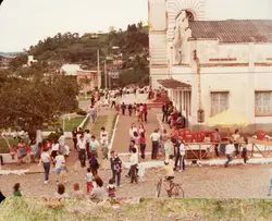 Uma cena ao ar livre em uma pequena cidade brasileira, mostrando uma praça ou pátio adjacente a uma igreja de arquitetura clássica. O ambiente sugere um evento comunitário ou festividade religiosa, com muitas pessoas circulando, crianças brincando e uma estrutura improvisada com guarda-sol amarelo e engradados de bebidas, típica de décadas passadas como os anos 80.