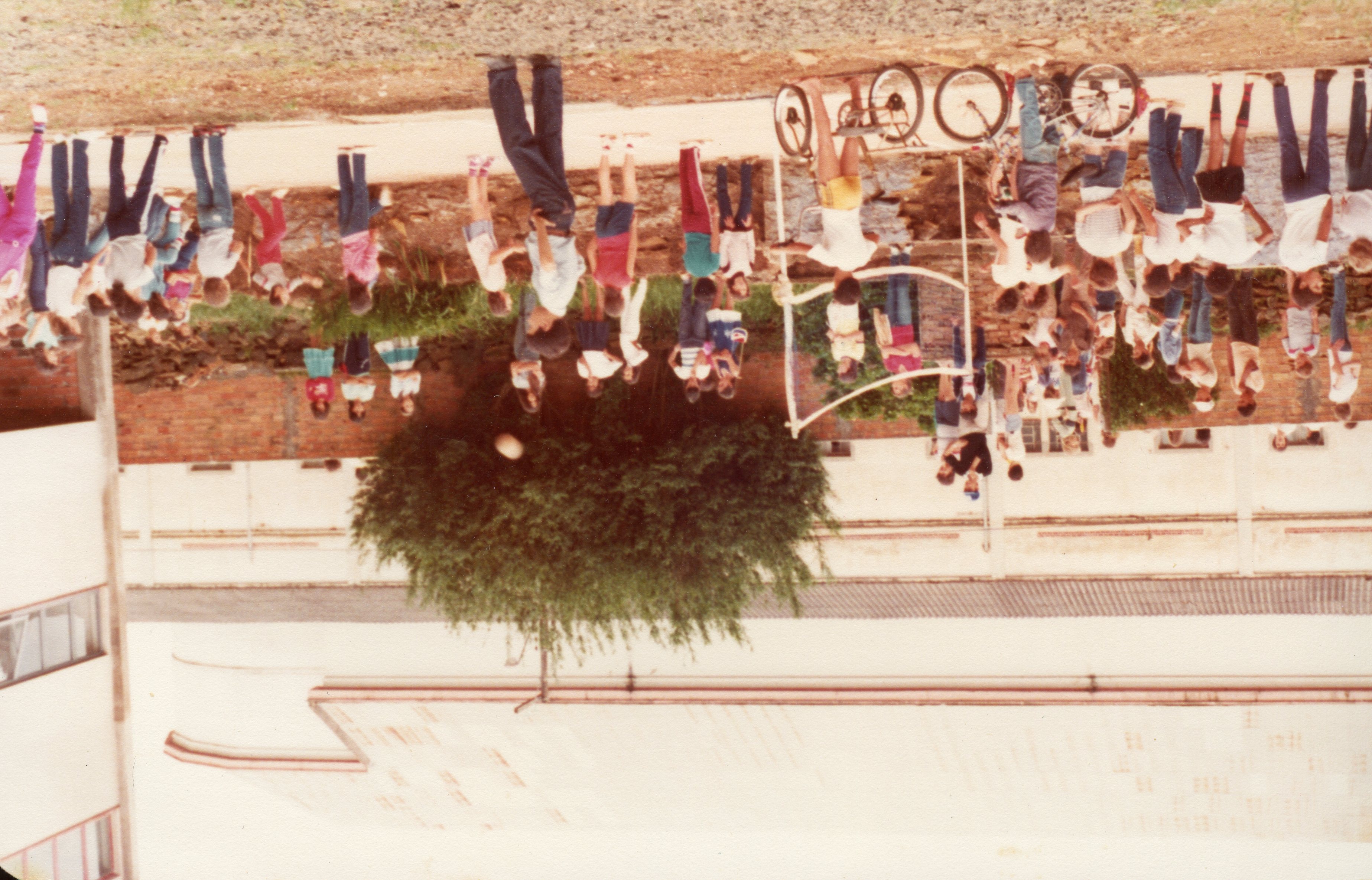 Uma fotografia colorida, orientada verticalmente, que captura um grupo de crianças e jovens reunidos em uma área externa, possivelmente um pátio escolar ou recreativo. A cena sugere uma atividade de lazer ou evento comunitário, com a presença de bicicletas e roupas típicas das décadas de 1980 ou 1990, refletindo um ambiente cotidiano de uma cidade brasileira.