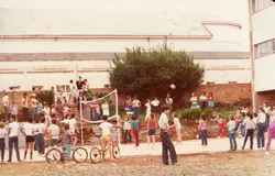 Uma fotografia colorida, orientada verticalmente, que captura um grupo de crianças e jovens reunidos em uma área externa, possivelmente um pátio escolar ou recreativo. A cena sugere uma atividade de lazer ou evento comunitário, com a presença de bicicletas e roupas típicas das décadas de 1980 ou 1990, refletindo um ambiente cotidiano de uma cidade brasileira.