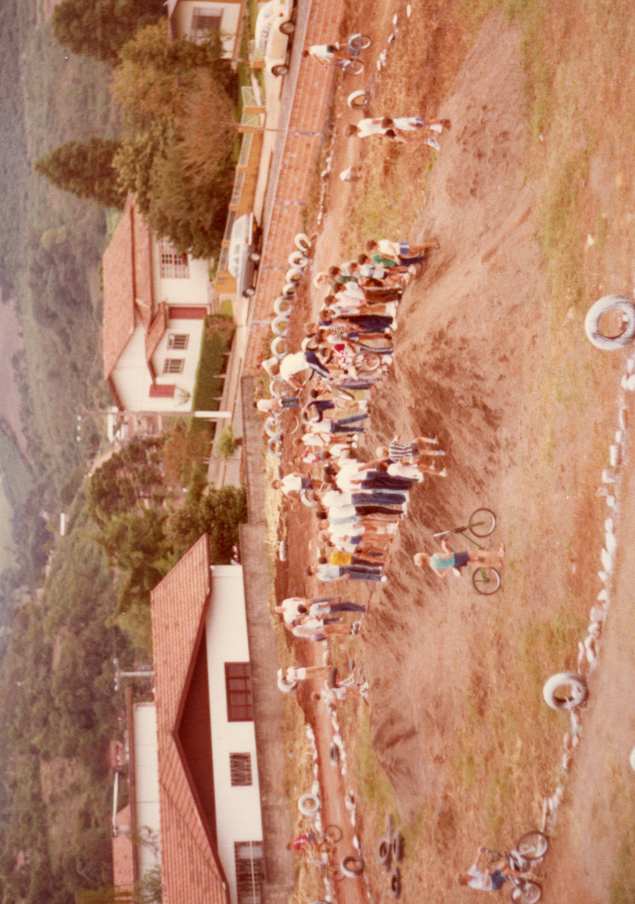 Uma fotografia aérea capturando um grupo de pessoas reunidas em um terreno de terra, possivelmente em uma área rural ou periurbana, com bicicletas espalhadas pelo local. A cena sugere um evento comunitário ou uma competição amadora de ciclismo, com pneus usados para delimitar o percurso, remetendo à estética e vestimentas típicas das décadas de 1980 ou 1990.