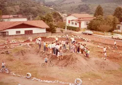 Uma fotografia aérea capturando um grupo de pessoas reunidas em um terreno de terra, possivelmente em uma área rural ou periurbana, com bicicletas espalhadas pelo local. A cena sugere um evento comunitário ou uma competição amadora de ciclismo, com pneus usados para delimitar o percurso, remetendo à estética e vestimentas típicas das décadas de 1980 ou 1990.