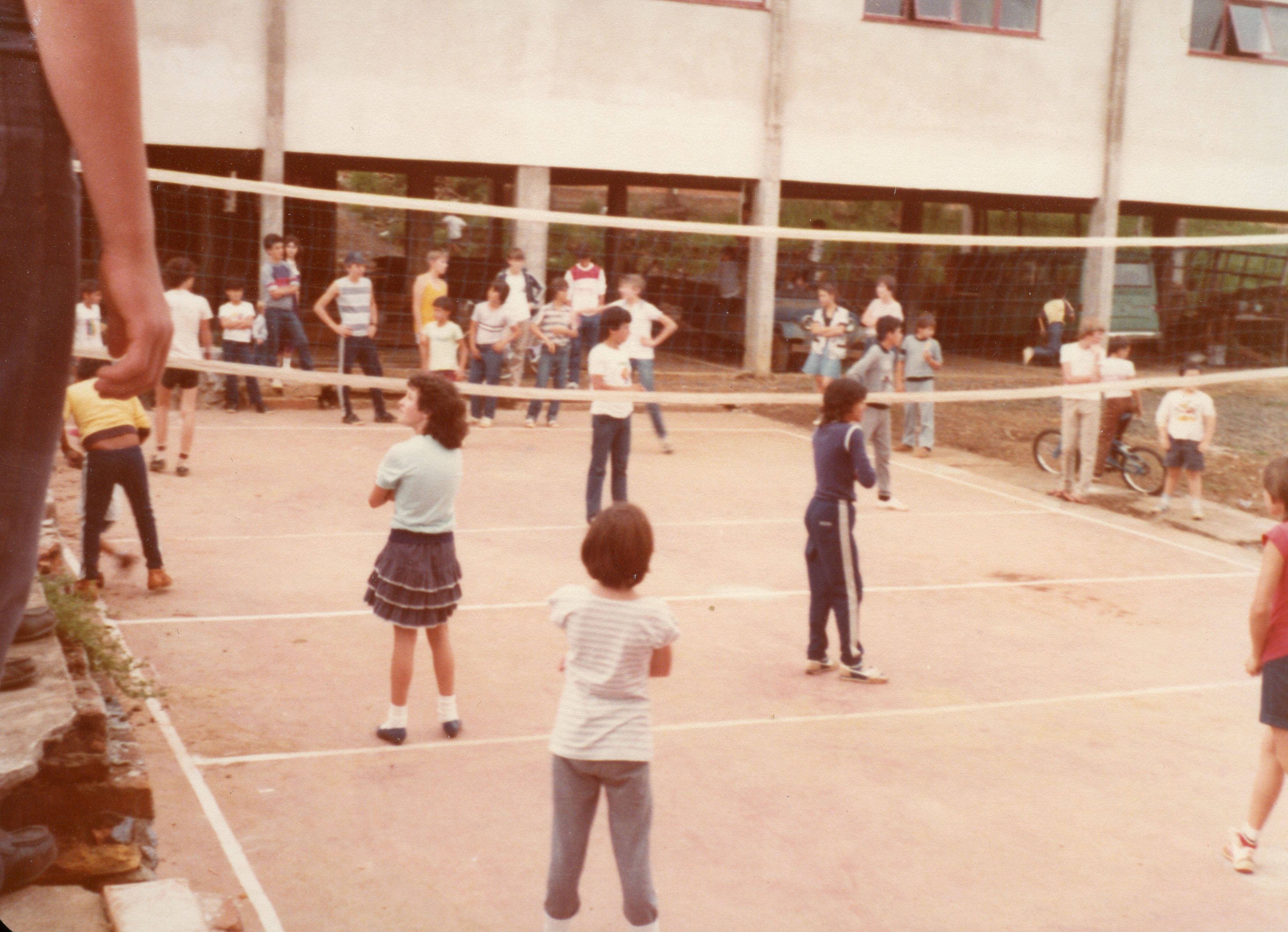 Uma fotografia que retrata um grupo de jovens e crianças jogando vôlei em uma quadra ao ar livre, possivelmente no pátio de uma escola. A vestimenta casual e o estilo da imagem sugerem um contexto escolar brasileiro entre o final da década de 1970 e início da década de 1980.