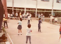 Uma fotografia que retrata um grupo de jovens e crianças jogando vôlei em uma quadra ao ar livre, possivelmente no pátio de uma escola. A vestimenta casual e o estilo da imagem sugerem um contexto escolar brasileiro entre o final da década de 1970 e início da década de 1980.