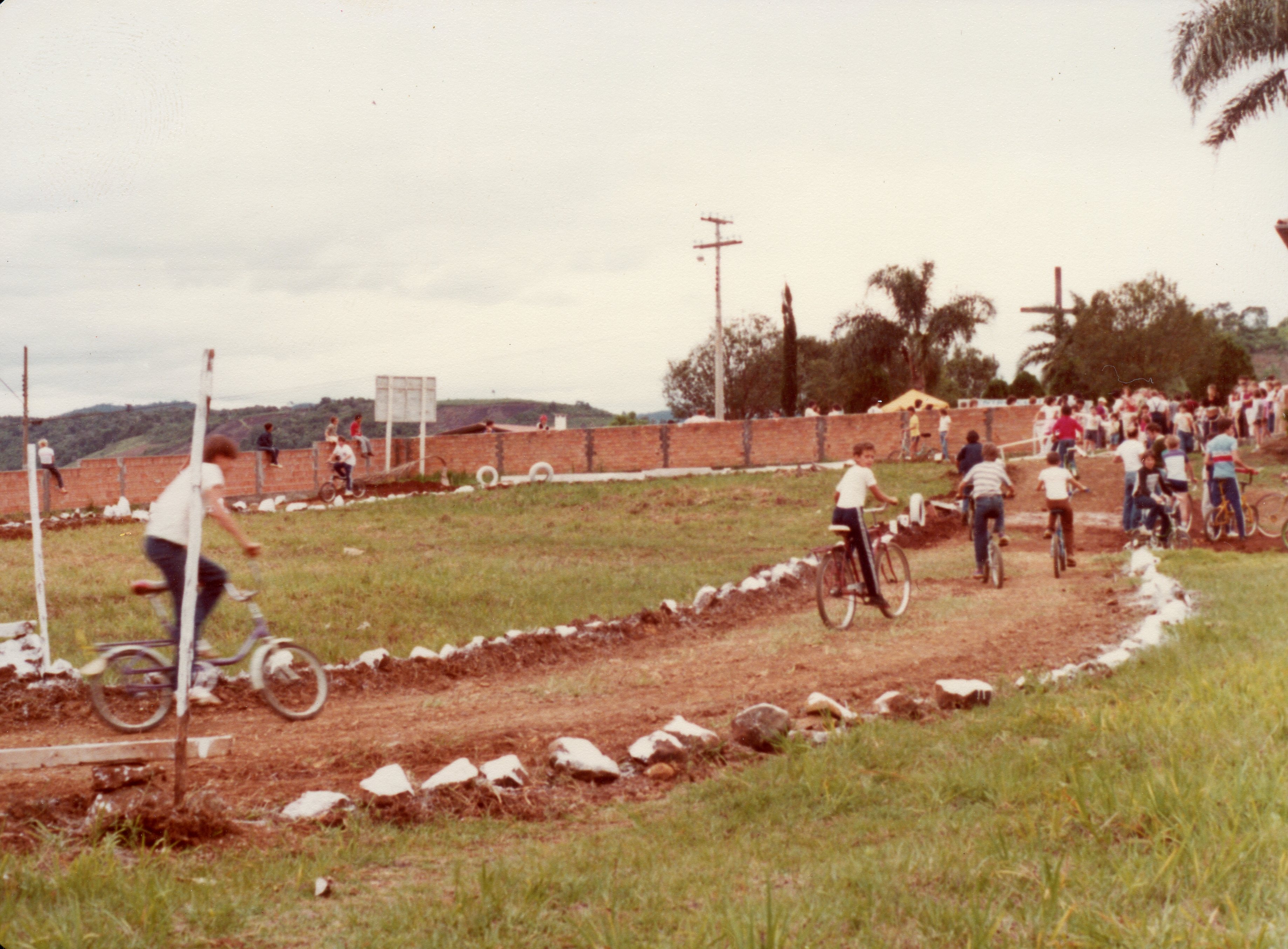 Uma cena ao ar livre mostrando um grupo de jovens participando de uma competição ou passeio de bicicleta em uma pista de terra improvisada. A imagem, provavelmente da década de 1980, apresenta um ambiente rural ou de periferia urbana com uma cerca de tijolos ao fundo e vegetação típica brasileira.