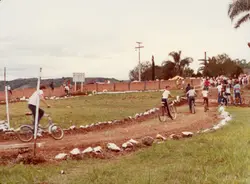 Uma cena ao ar livre mostrando um grupo de jovens participando de uma competição ou passeio de bicicleta em uma pista de terra improvisada. A imagem, provavelmente da década de 1980, apresenta um ambiente rural ou de periferia urbana com uma cerca de tijolos ao fundo e vegetação típica brasileira.