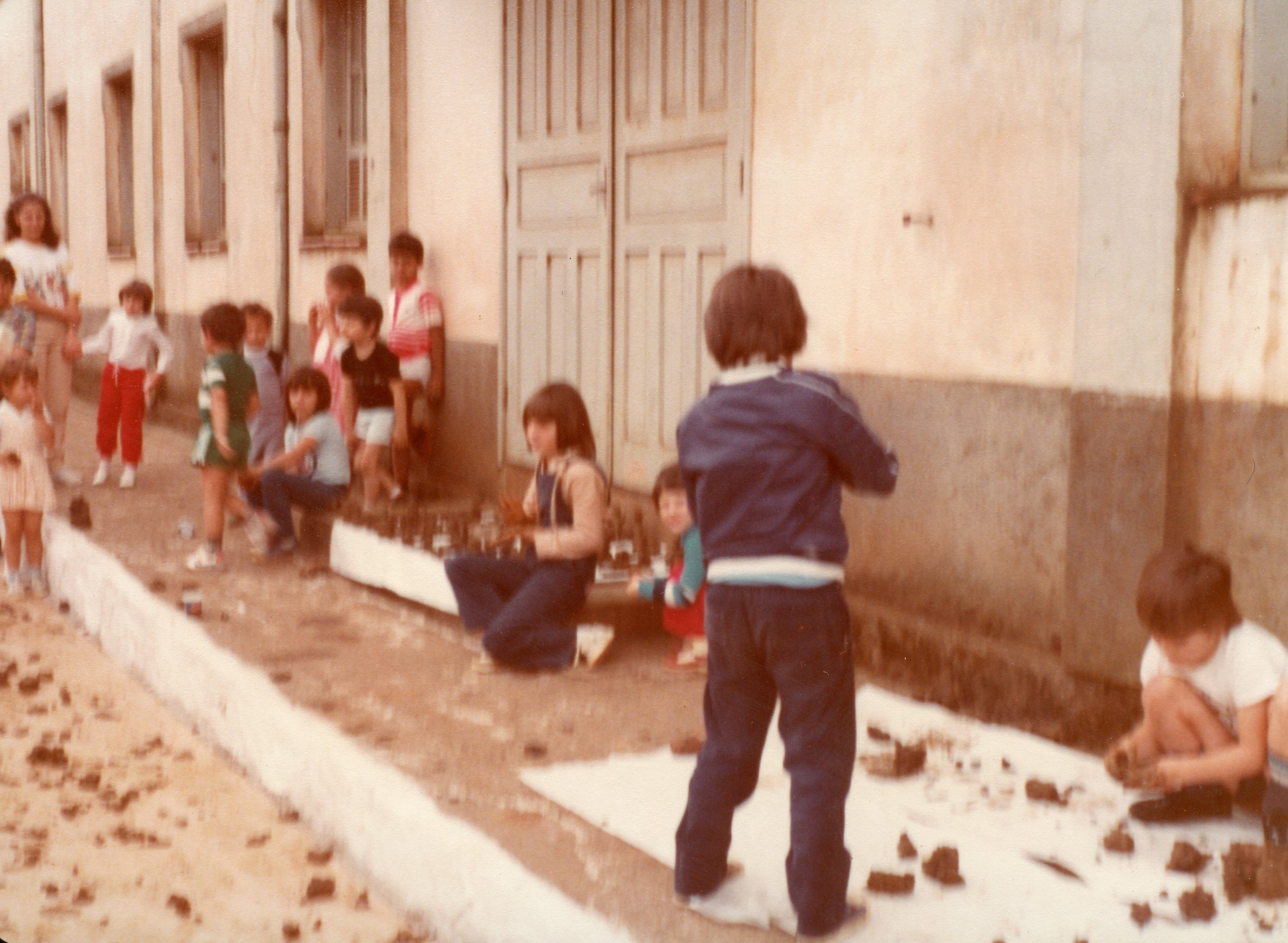 Uma fotografia colorida que retrata um grupo de crianças brincando ao ar livre em frente a uma construção de alvenaria, possivelmente uma escola ou prédio público. As crianças estão sentadas ou agachadas no chão, manipulando objetos ou terra, sugerindo uma atividade recreativa ou escolar típica das décadas de 1980 ou 1990.