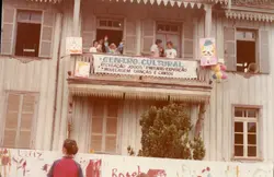 Uma fotografia vertical mostrando a fachada de uma casa de madeira tradicional com arquitetura típica de colonização europeia no sul do Brasil. O local está decorado com uma faixa indicando um 'Centro Cultural' e balões, sugerindo um evento recreativo ou infantil, possivelmente ocorrido entre as décadas de 1980 e 1990.